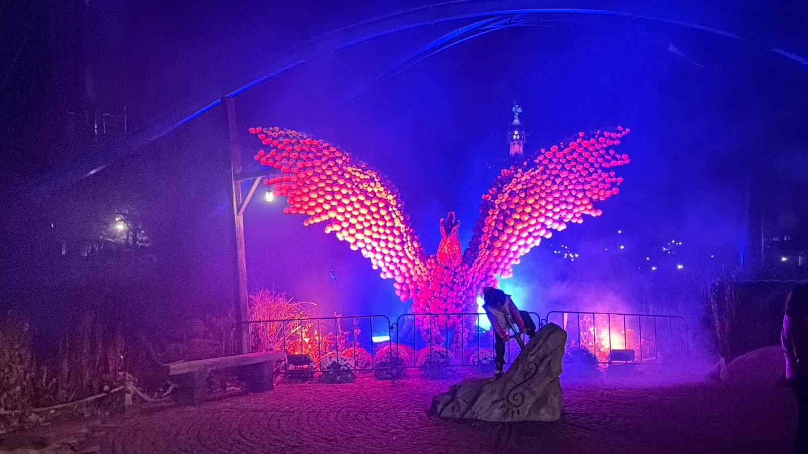 Riesiger, aus tausenden leuchtenden Kürbissen geformter Phönix mit ausgebreiteten Flügeln bei Nacht, davor ein Darsteller mit Vorschlaghammer